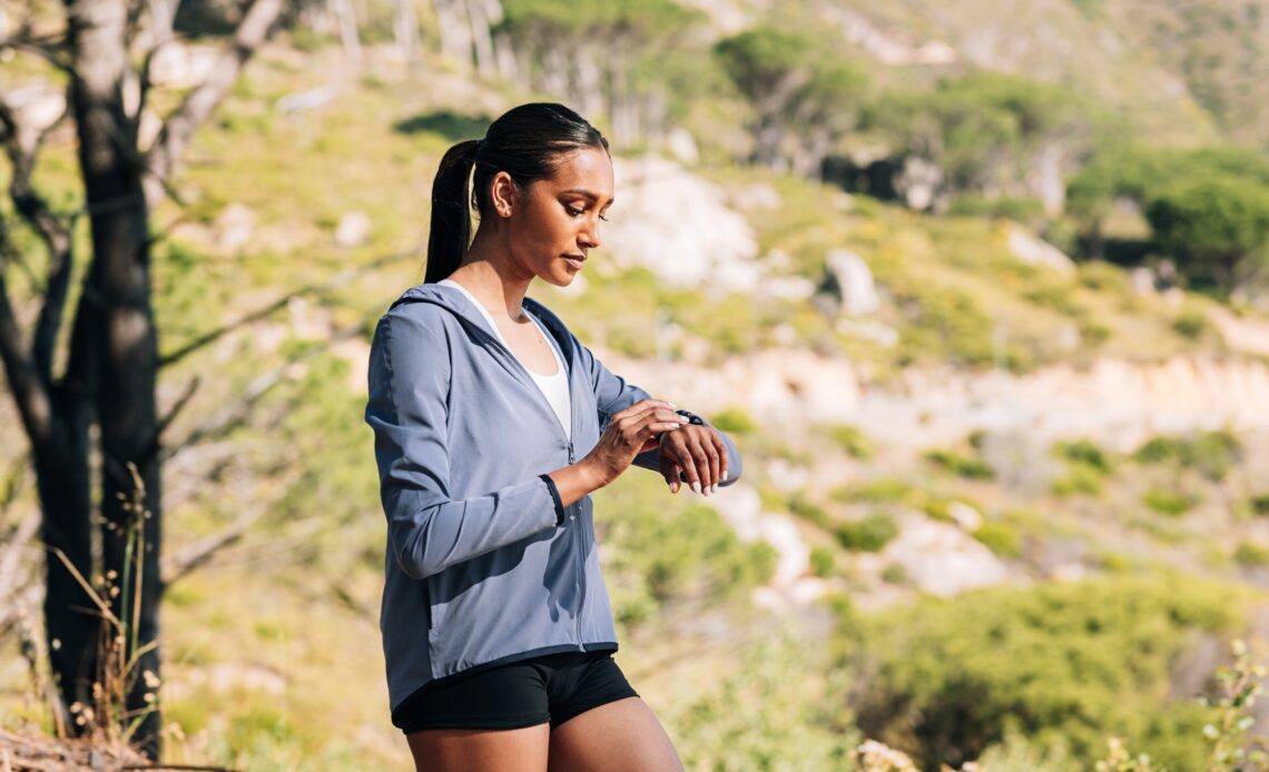 Young slim female in fitness attire walking in a natural park and looking at her smartwatches